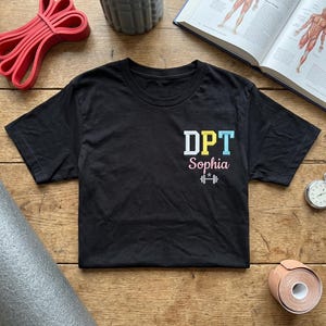 May include: Black t-shirt featuring the text "DPT Sophia" in white, yellow, and blue, and a barbell icon. The shirt is on a wooden surface, with exercise equipment and a book visible in the background.