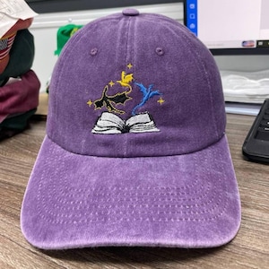 May include: A purple baseball cap featuring an embroidered design of an open book with a black dragon, a yellow dragon, and a blue dragon emerging from the pages, accented by small yellow stars above the book on the front of the cap.
