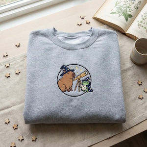 May include: A gray sweatshirt with an embroidered design featuring a capybara and a frog, both wearing wizard hats and looking through a telescope. The design is set within a circular frame with stars and a crescent moon.