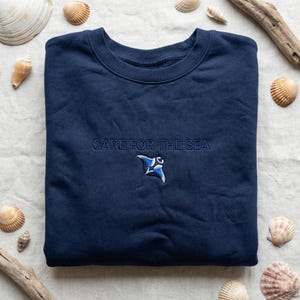 Könnte beinhalten: Marineblaues Sweatshirt mit dem gestickten Schriftzug "CARE FOR THE SEA" über einem blau-weißen Mantarochen-Design. Das Sweatshirt ist von Muscheln und Treibholz umgeben.