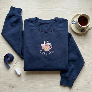 May include: A navy blue sweatshirt featuring an embroidered design of a pink teapot, two white ducks, and the words "Cozy Tea." A cup of tea on a saucer is visible. A spool of thread and a blue ribbon are also present.