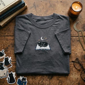 May include: Dark gray t-shirt featuring an embroidered design of a sleeping black cat on an open book, with a crescent moon and stars above. The shirt is a crew neck style and is displayed on a wooden surface.