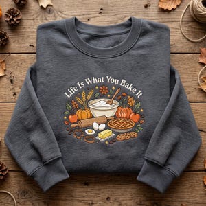 May include: A charcoal gray crewneck sweatshirt featuring a central graphic with the text "Life Is What You Bake It" surrounded by illustrations of a mixing bowl, rolling pin, pie, pumpkins, apples, wheat, eggs, butter, and cookies.