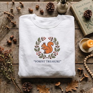 May include: A white crewneck sweatshirt featuring an embroidered design of an orange squirrel centered within a circular wreath of green oak leaves and red berries, with the text "FOREST TREASURE" printed in dark blue lettering below.