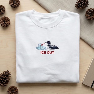 Peut inclure: Sweat-shirt blanc avec un motif brodé représentant un huard, un glaçon et les mots "ICE OUT" en rouge. Le huard est noir et blanc, et le glaçon est bleu. Le sweat-shirt est en matière douce.