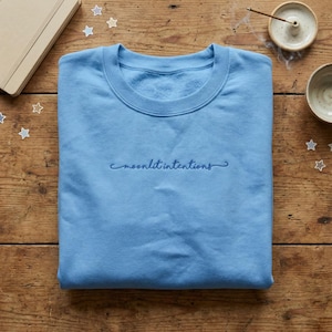 May include: A light blue sweatshirt with the embroidered text "moonlit intentions" in a matching blue thread. The crew neck sweatshirt is on a wooden surface with a notebook, stars, incense, and a small bowl.