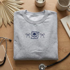 May include: A grey sweatshirt with a navy blue ultrasound machine design and bow accents. The sweatshirt is made of a soft material. Suitable for medical professionals or those in the healthcare field.