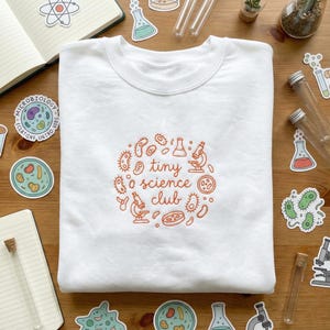 May include: White crewneck sweatshirt with an orange embroidered design. The design features the words "tiny science club" surrounded by illustrations of microbes, microscopes, and lab equipment. The sweatshirt is surrounded by science-themed stickers and lab equipment.