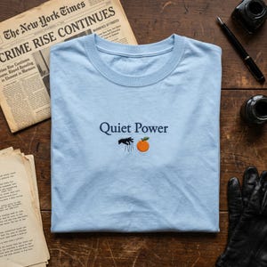 May include: A light blue t-shirt with the embroidered text "Quiet Power" above a graphic of a black figure and an orange. The shirt is folded on a wooden surface, with a vintage newspaper and inkwells in the background.