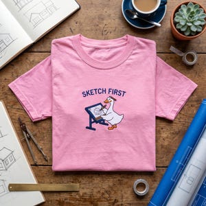 May include: A light pink t-shirt featuring an embroidered graphic of a white goose standing at a drafting table with a pencil in its beak and one on its head. The text SKETCH FIRST is embroidered in blue above the goose, which is drawing a house.