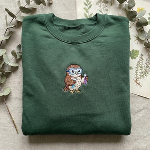 May include: Dark green sweatshirt featuring an embroidered owl wearing goggles and holding a test tube. The owl is brown, white, and tan. The sweatshirt is folded on a neutral surface.