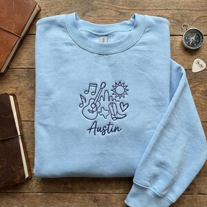 May include: A light blue sweatshirt with a crew neck. The front features an embroidered design of a guitar, musical notes, a cityscape, a sun, cowboy boots, and the word "Austin" in a script font. The sweatshirt is on a wooden surface.