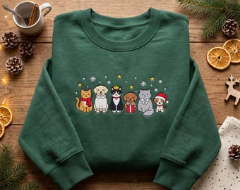 May include: A forest green crewneck sweatshirt featuring a central graphic of six cartoon animals including cats and dogs wearing winter accessories like scarves, a Santa hat, and a crown, surrounded by small white snowflakes and yellow stars.