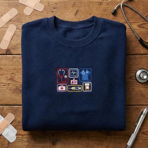 Könnte beinhalten: Marineblaues Sweatshirt mit gestickten medizinischen Symbolen, darunter ein Stethoskop, ein EKG, eine OP-Kleidung und eine Pillenflasche. Das Design ist in Rot, Weiß, Gelb und Blau gehalten. Das Sweatshirt liegt auf einer Holzoberfläche.
