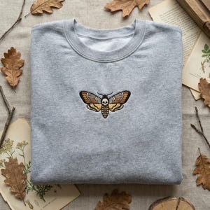 May include: A gray sweatshirt with a detailed embroidered moth design. The moth features brown, yellow, and black wings with a skull detail. The sweatshirt is folded on a neutral-toned surface with dried leaves and botanical illustrations.