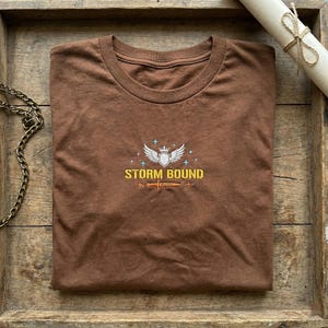 May include: A brown crew neck t-shirt featuring the embroidered text "STORM BOUND" above a sword and wing emblem. The shirt is neatly folded on a wooden surface, with a decorative chain and scroll in the background.