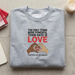 Könnte beinhalten: Ein graues Sweatshirt mit dem Text "THE ONLY THING MORE POWERFUL THAN HATE IS LOVE" in Schwarz und Rot. Das Wort "LOVE" ist in großen roten Buchstaben geschrieben. Darunter bilden zwei Hände ein Herz, mit den Worten "TOGETHER, WE ARE AMERICA".