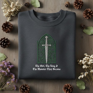 May include: Dark gray sweatshirt with an embroidered design of a sword inside a green arched frame. The text "The Girl, The King & The Monster They Became" is below the design. The sweatshirt is surrounded by decorative elements.