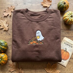 Peut inclure: Un sweat-shirt à col rond marron foncé avec une broderie représentant un fantôme blanc tenant un arrosoir jaune, s'occupant d'un petit groupe de citrouilles orange et de vignes vertes. Le sweat-shirt possède un col côtelé et des manches longues.