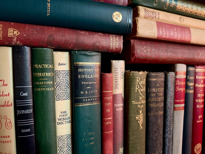 May include: A collection of antique books with various colors and titles. The spines are visible, showcasing titles like "Practical Pediatrics" and "History of England." The books are stacked and arranged, creating a vintage aesthetic.