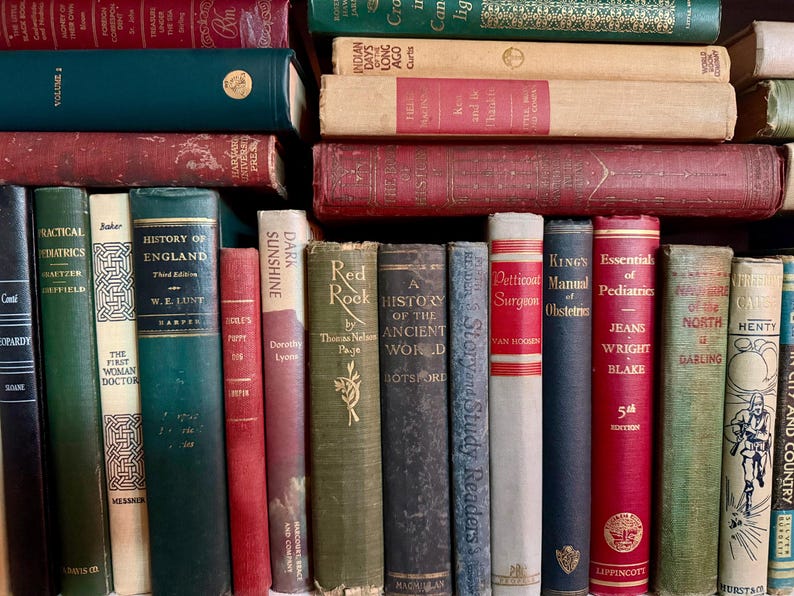 May include: A collection of antique books with various colors and sizes. The spines display titles like "History of England" and "Essentials of Pediatrics." The books are stacked and arranged, creating a vintage library aesthetic.