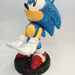 Puede incluir: Una figura de Sonic the Hedgehog, cubierta de brillantes pedrería azul. La figura tiene guantes blancos, zapatos rojos con purpurina y una base negra. La cara del personaje es amarilla y blanca.