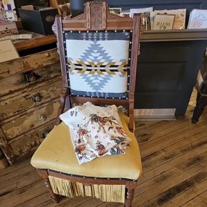 May include: An antique wooden chair with a yellow leather seat and fringe trim. The backrest has a geometric patterned fabric in grey, black, and gold. A white t-shirt with a cowboy graphic is draped over the seat.