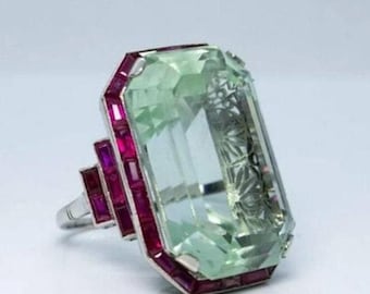 May include: A large, emerald green gemstone ring with a rectangular cut. The ring is set in silver and has a border of small, red ruby stones.