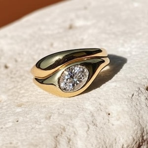 Puede incluir: Un anillo de oro con un diseño único y fluido. Presenta un diamante ovalado engastado en una banda de oro pulido. La banda tiene una forma suave y curvada, creando un aspecto moderno y elegante. El anillo se muestra sobre una superficie texturizada de color blanco roto.
