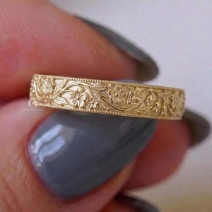 May include: A gold band ring featuring an intricate, continuous embossed floral pattern with small flowers and leaves. The metal surface has a polished finish, and the edges of the band are detailed with a fine, beaded milgrain texture.