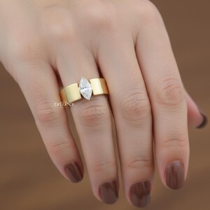Puede incluir: Un anillo de oro con un diamante talla marquesa. El anillo tiene una banda ancha con un dise&ntilde;o plano y rectangular a cada lado del diamante. El anillo est&aacute; en una mano con esmalte de u&ntilde;as marr&oacute;n. La palabra "OPULENCE" es visible.