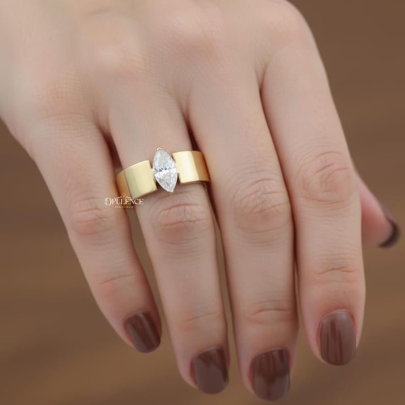 Puede incluir: Un anillo de oro con un diamante talla marquesa. El anillo tiene una banda ancha con un dise&ntilde;o plano y rectangular a cada lado del diamante. El anillo est&aacute; en una mano con esmalte de u&ntilde;as marr&oacute;n. La palabra "OPULENCE" es visible.