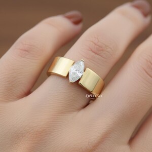 Puede incluir: Un anillo de oro con un diamante talla marquesa. El anillo tiene un dise&ntilde;o moderno con una banda ancha y plana. El diamante est&aacute; engastado en el centro, entre dos secciones de oro. El anillo est&aacute; en el dedo de una persona.