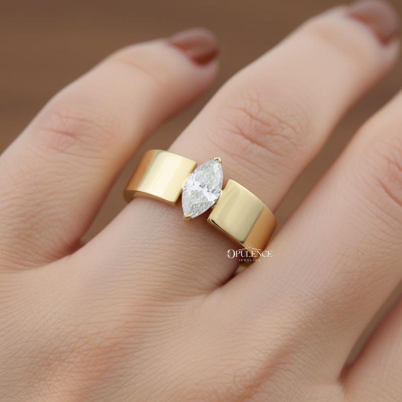 Puede incluir: Un anillo de oro con un diamante talla marquesa. El anillo tiene un dise&ntilde;o moderno con una banda ancha y plana. El diamante est&aacute; engastado en el centro, entre dos secciones de oro. El anillo est&aacute; en el dedo de una persona.