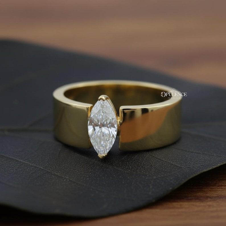 Puede incluir: Un anillo de oro con un diamante talla marquesa. El diamante est&aacute; engastado verticalmente, y la banda es de oro pulido grueso. El anillo se muestra sobre una superficie oscura.
