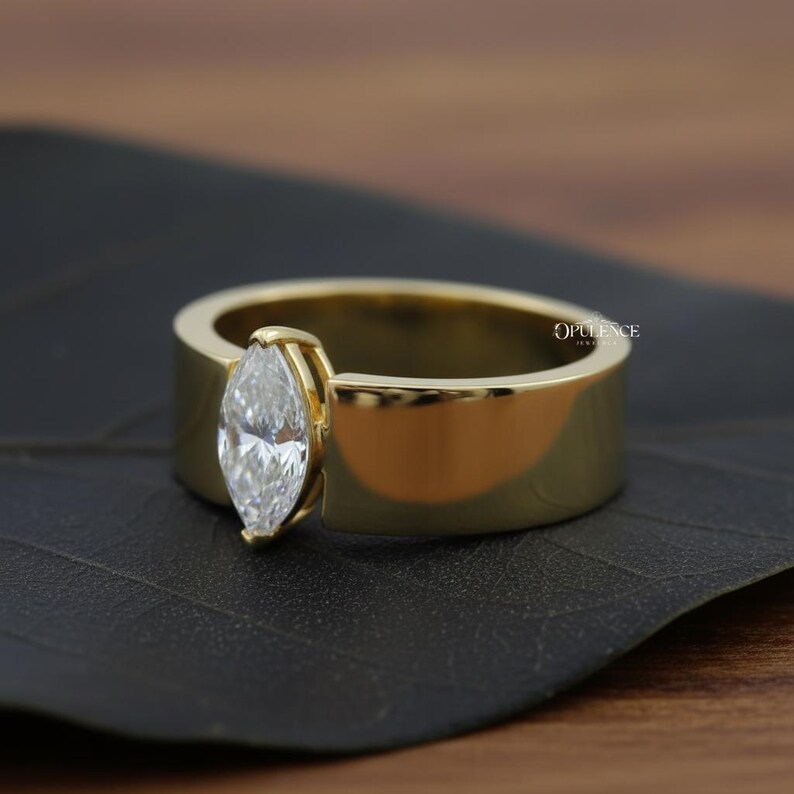 Puede incluir: Un anillo dorado con un diamante talla marquesa. El anillo tiene una banda ancha y un engaste biselado que sujeta el diamante. El anillo se muestra sobre una superficie oscura. La palabra "OPULENCE" es visible en el anillo.