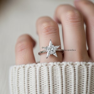 May include: A silver ring featuring a star-shaped, clear gemstone. The ring's design includes a thin band and a five-pointed star setting. The ring is shown on a finger, with a white sweater in the background.