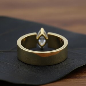 Puede incluir: Un anillo dorado con una gema de talla marquesa. El anillo tiene una banda gruesa y un engaste que sujeta la gema. El anillo se muestra sobre una superficie oscura, resaltando su acabado pulido. El anillo es una joya.