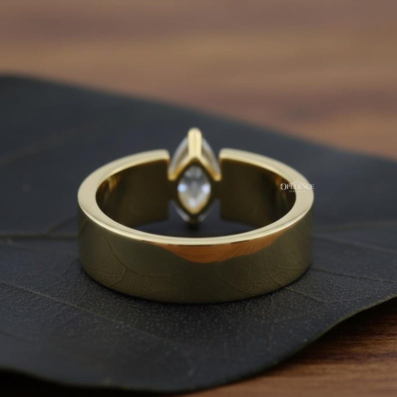 Puede incluir: Un anillo dorado con una gema de talla marquesa. El anillo tiene una banda gruesa y un engaste que sujeta la gema. El anillo se muestra sobre una superficie oscura, resaltando su acabado pulido. El anillo es una joya.