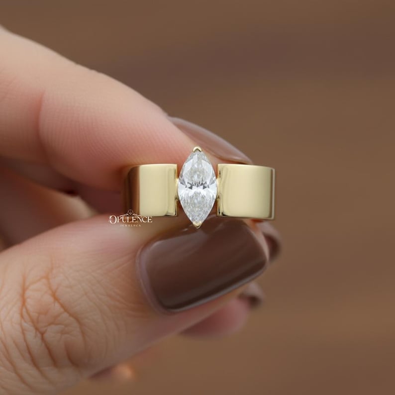Puede incluir: Un anillo de oro con un diamante talla marquesa. El diamante est&aacute; engastado entre dos bandas rectangulares de oro. El anillo est&aacute; sostenido por una mano con esmalte de u&ntilde;as marr&oacute;n. La palabra "OPULENCE" est&aacute; impresa en el anillo.