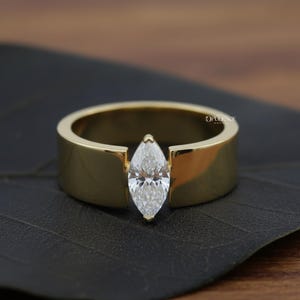 Puede incluir: Un anillo dorado con un diamante talla marquesa. El anillo tiene una banda ancha y plana con un engaste en canal que sujeta el diamante. El anillo se muestra sobre una superficie oscura.