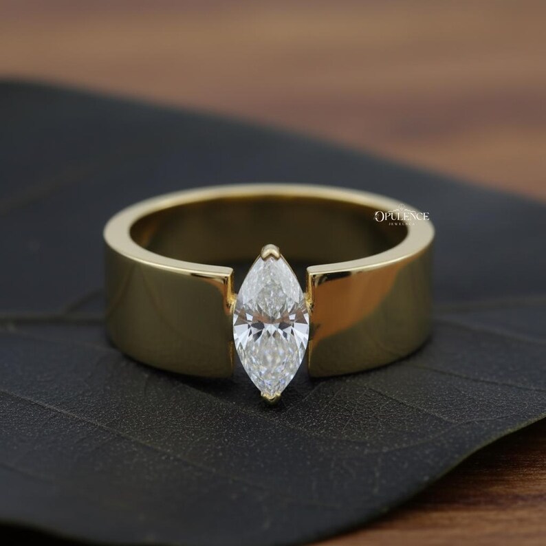 Puede incluir: Un anillo dorado con un diamante talla marquesa. El anillo tiene una banda ancha y plana con un engaste en canal que sujeta el diamante. El anillo se muestra sobre una superficie oscura.
