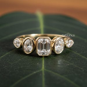 May include: A gold ring featuring an emerald-cut diamond center stone, flanked by oval and pear-shaped diamonds. The ring is set on a green leaf, with the word "OPULENCE" visible.