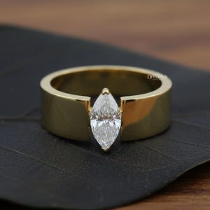 Puede incluir: Un anillo de oro con un diamante talla marquesa. El diamante est&aacute; engastado entre la banda gruesa y plana del anillo. El anillo se muestra sobre una superficie oscura, resaltando el oro pulido y el brillo del diamante.