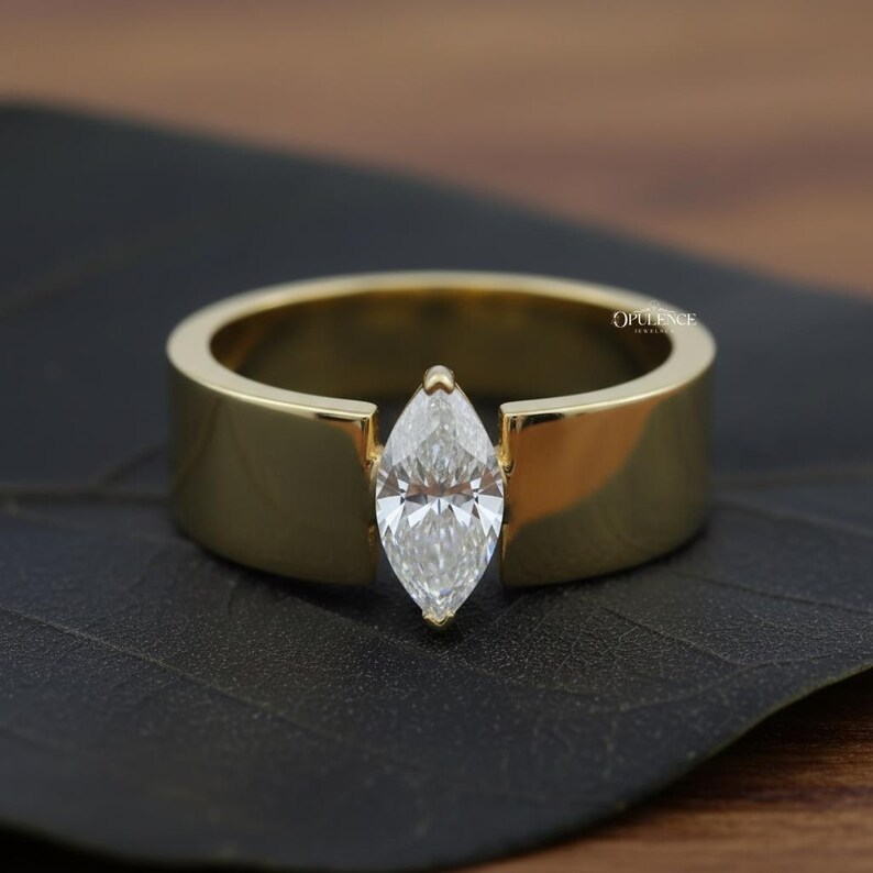 Puede incluir: Un anillo de oro con un diamante talla marquesa. El diamante est&aacute; engastado entre la banda gruesa y plana del anillo. El anillo se muestra sobre una superficie oscura, resaltando el oro pulido y el brillo del diamante.