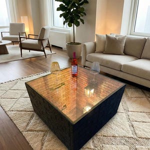 May include: A modern living room scene featuring a black square coffee table with a mirrored top reflecting a brick pattern. A bottle of rosé wine and two wine glasses sit on the table. The room includes a beige sofa, chairs, and a large potted plant.