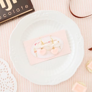 Könnte beinhalten: Hellrosa herzförmige Ohrringe mit goldfarbenen Haken und einer weißen Schleife. Die Ohrringe werden auf einer rosa Karte mit dem Text "Dessert Collection" präsentiert. Sie liegen auf einem weißen Teller.