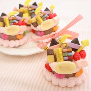 Könnte beinhalten: Miniatur-Dekokuchen mit rosa Zuckerguss, Schokoladenschichten und bunten Belägen wie Erdbeeren, Bananen und Süßigkeiten. Die Kuchen sind mit Bändern und einem Schlüsselanhänger verziert. Die Kuchen stehen auf einem weißen Teller und einem rosa gestreiften Hintergrund.