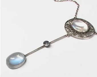 Puede incluir: Un collar de tono plateado con un colgante circular que presenta una piedra de luna ovalada central, rodeada de intrincados adornos metálicos. Una barra vertical desciende sosteniendo una pequeña piedra redonda y una piedra de luna ovalada.