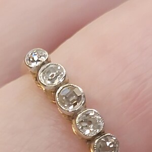 May include: A gold ring featuring five round, clear, faceted stones set in a row. Each stone is held in a circular metal setting, creating a continuous line of sparkle across the band. The design highlights the clarity and brilliance of the stones.